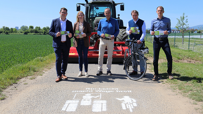 Bürgermeister Sebastian Wysocki, Erste Kreisbeigeordnete Birgit Weckler, Kreislandwirt Michael Schneller, ADFC-Sprecher Walter Baumann und Julian Herzel, Nahmobilitätskoordinator des Wetteraukreises. Sie stehen auf einem Feldweg, auf dem Weg ein Piktogramm "Rücksicht macht Wege breit.