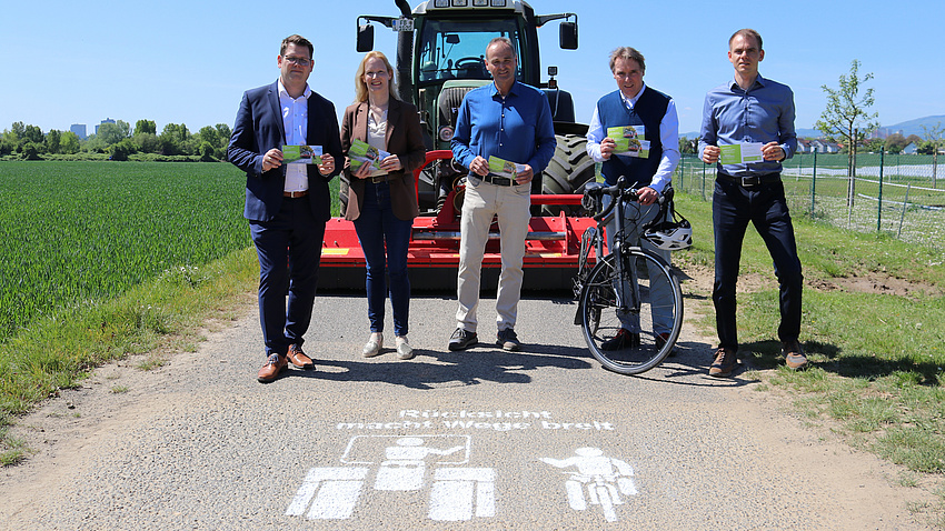 Bürgermeister Sebastian Wysocki, Erste Kreisbeigeordnete Birgit Weckler, Kreislandwirt Michael Schneller, ADFC-Sprecher Walter Baumann und Julian Herzel, Nahmobilitätskoordinator des Wetteraukreises. Sie stehen auf einem Feldweg, auf dem Weg ein Piktogramm "Rücksicht macht Wege breit.