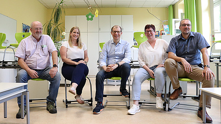 Fünf Erwachsene sitzen auf kleinen Stühlen in einem Klassenzimmer einer Grundschule.