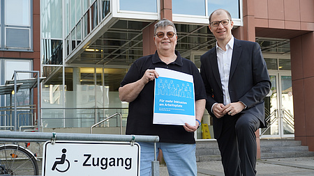 Zwie Menschen stehen an einem Schild, das den barrierefreien Zugang ausschildert.