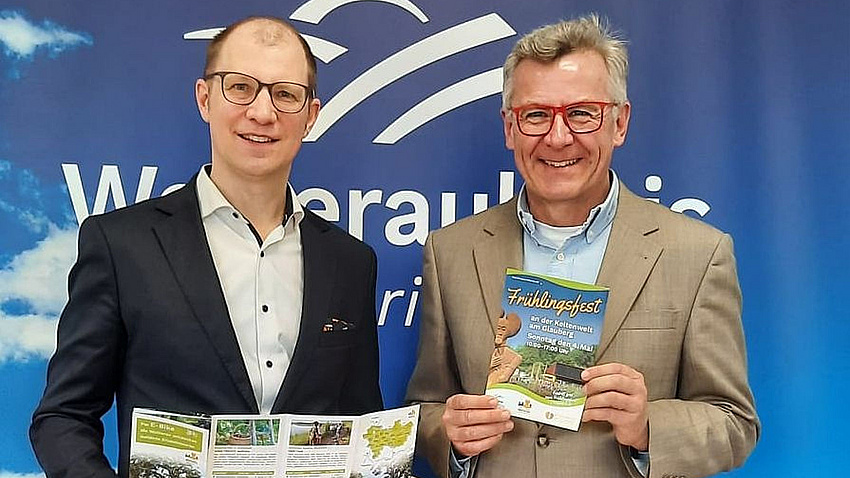 Zwei Männer stehen vor einem blau-weißen Hintergrund mit der Aufschrift Wetteraukreis und halten Flyer in den Händen.