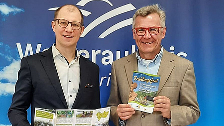 Zwei Männer stehen vor einem blau-weißen Hintergrund mit der Aufschrift Wetteraukreis und halten Flyer in den Händen.