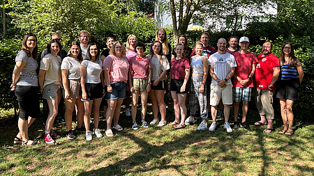 Eine Gruppe junger Leute steht in Freizeitkleidung auf einer Wiese. Alle lächeln in die Kamera.