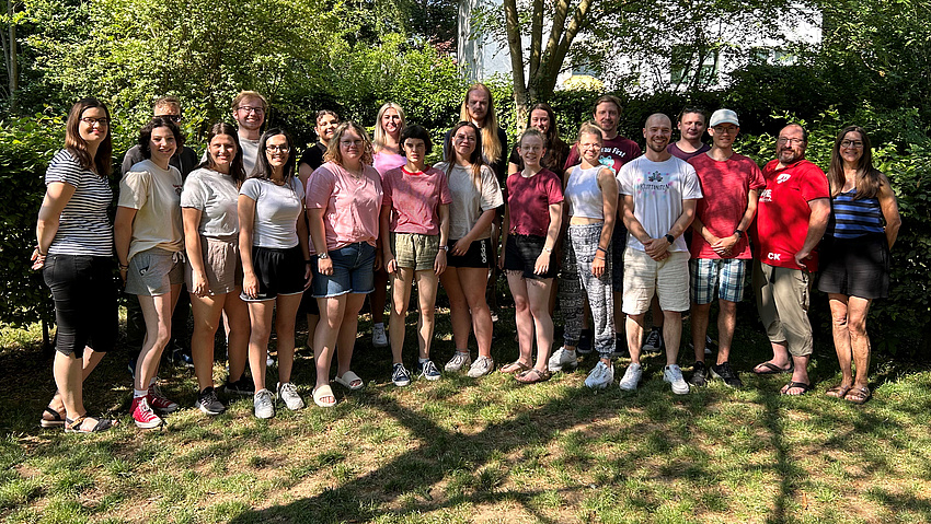 Eine Gruppe junger Leute steht in Freizeitkleidung auf einer Wiese. Alle lächeln in die Kamera.