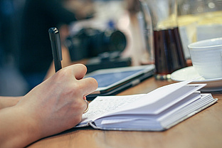 Foto einer Hand, die einen Stift hält, und in ein Notizbuch schreibt.
