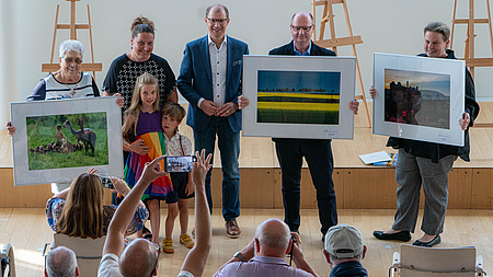 Fünf Erwachsene und zwei Kinder. Drei Personen halten ein großformatiges Bild in der Hand. Im Hintergrund drei Staffeleien.