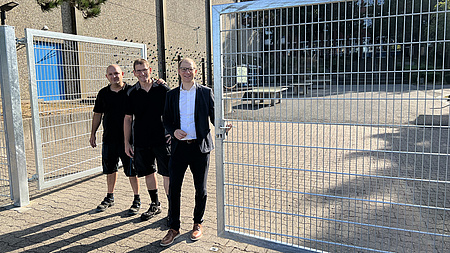 Drei Männer stehen vor einem Zaun, im Hintergrund ist ein Schulhof mit Tischtennisplatte zu sehen.