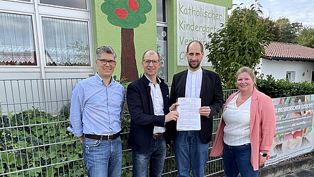Eine Frau und drei Männer stehen vor einem Haus. Zwei Männer halten ein Schriftstück in der Hand.