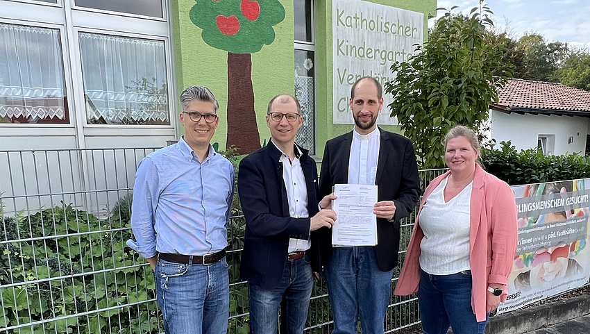 Eine Frau und drei Männer stehen vor einem Haus. Zwei Männer halten ein Schriftstück in der Hand.