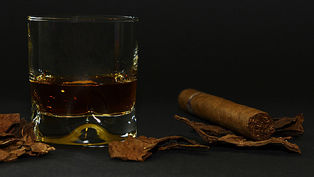 Ein gefülltes Whiskey-Glas und ein Zigarillo liegen auf einer dunklen Tischoberfläche nebeneinander.