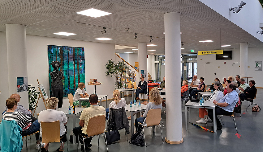 An kleinen Tischen, die im Halbkreis aufgestellt sind sitzen mehrere Personen und höen einer Frau zu, die aus einem Buch liest.