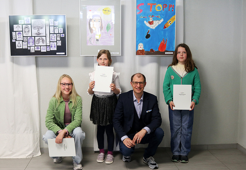 Drei Mädchen und ein Mann. Die Mädchen halten Urkunden in der Hand. Der Mann und eines der Mädchen in der Hocke. Im Hintergrund hängen Bilder an der Wand.