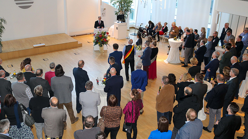 Ein Saal von oben. Unten stehen viele Menschen und hören einem Mann zu, der an einem Rednerpult steht.