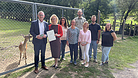 Eine Gruppe aus acht Personen steht im Freien auf einer Wiese vor einem mannshohen Drahtzaun. Hinter dem Zaun steht ein Wildtier. Eine Frau der Gruppe hält eine Dokumentenmappe in Händen.