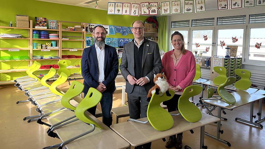 Zwei Männer und eine Frau stehen in einem Klassenraum. Vor und neben ihnen befinden sich mehrere Schultische und Stühle und im Hintergrund stehen Regale.