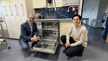 Eine Frau und ein Mann knien sich in einem Laborraum neben eine geöffnete Spülmaschine.