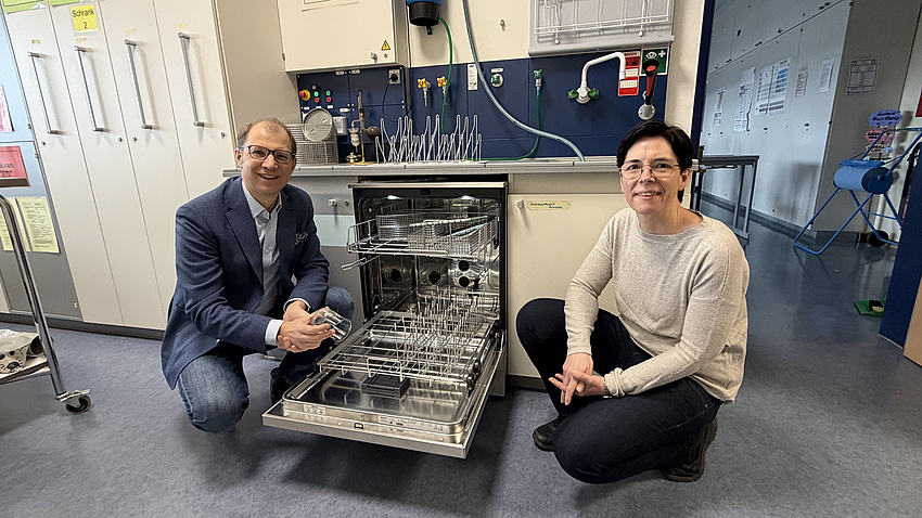 Eine Frau und ein Mann knien sich in einem Laborraum neben eine geöffnete Spülmaschine.