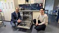 Eine Frau und ein Mann knien sich in einem Laborraum neben eine geöffnete Spülmaschine.