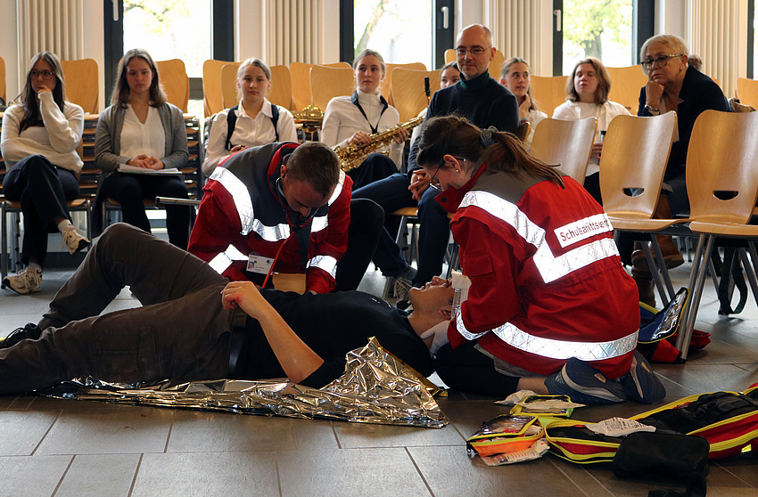 Ein junger Mann liegt auf dem Boden und wird von zwei Personen rettungsdienstlich versorgt. Im Hintergrund Personen, die zusehen.