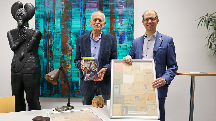 Zwei Herren stehen vor einem großen Gemälde, einer hält ein Buch in der Hand, der andere einen Bilderrahmen.