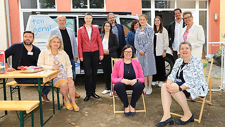 Mehrere Personen stehen oder sitzen auf Bänken und in Sonnenstühlen vor einem Van, der vor einem Gebäude steht.