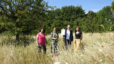 Eine Gruppe von Menschen steht in einem Feld, im Hintergrund sind Bäume zu sehen. Sie präsentieren ein Schild.